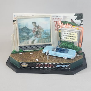 May include: A miniature diorama of a 1961 Dodge Dart Phoenix car in front of a movie theatre marquee with the text "See Elvis Sing, Dance and Romance in Blue Hawaii." The diorama is on a black base with a film strip design and the text "1961 Dodge Dart Phoenix Blue Hawaii."