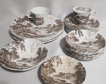 May include: A collection of vintage brown and white ceramic dishes. The set includes teacups, saucers, bowls, and plates, all featuring a detailed landscape scene. The dishes are arranged on a white surface, showcasing the complete set.
