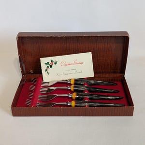 May include: A vintage boxed set of forks with black handles, yellow accents, and silver tines. The forks are arranged in a red velvet-lined box with a greeting card that reads "Christmas Greetings."