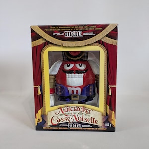 May include: A red M&M's Nutcracker candy dispenser in a box with a red and gold stage curtain design. The box reads "Nutcracker Casse-Noisette" and "Chocolate Candy Dispenser - Distributeur de bonbons au chocolat".