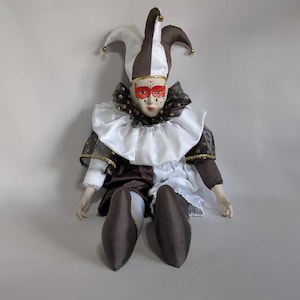 May include: A seated jester doll with a harlequin costume in white, brown, and black. The doll has a white face with red eye mask, red lips, and a brown and white jester hat with gold accents. The doll's outfit includes a ruffled collar.