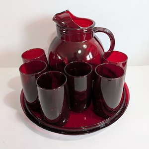 May include: A vintage ruby red glass drinkware set. The set includes a jug with a handle and six matching tumblers, all on a round tray. The glassware has a glossy finish, reflecting light. The set is ideal for serving drinks.