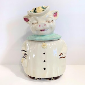 Puede incluir: Un tarro de galletas de cerámica con forma de cerdo con un diseño caprichoso. El cerdo lleva un sombrero con flores amarillas y un cuello azul claro. El tarro tiene un cuerpo blanco con botones negros y un pequeño adorno floral.