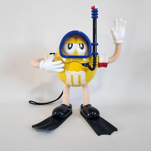 May include: A yellow M&M character dressed as a scuba diver with a blue diving mask, snorkel, and flippers. The character is waving with one hand and has a red and white buoy attached to the snorkel.