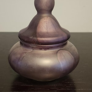 Puede incluir: Un pequeño tarro de vidrio morado redondo con tapa. El tarro tiene una superficie lisa y brillante y un acabado ligeramente iridiscente.