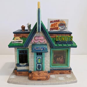 Puede incluir: Una tienda de donuts de cerámica en miniatura con fachada de ladrillo, adornos verdes y un letrero que dice "Patty's". La tienda presenta una "Walk-Up Window" y un letrero de "Delicious Hot Coffee". También es visible un letrero de "Donuts".