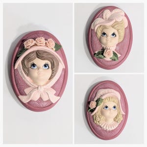 May include: Three oval wall plaques with a vintage lady design. Each plaque has a mauve border and a textured background. The ladies have blue eyes, rosy cheeks, and wear hats with flowers and feathers. A charming, decorative set for the home.