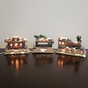 以下が含まれることがあります： 3両編成の照明付きセラミック製クリスマス列車セット。機関車は赤、緑、黒で、金色のベルが付いています。真ん中の車両は緑と茶色で、側面に「サンタの急行」と書かれています。最後の車両は茶色で、白い屋根と赤と緑の装飾が施されています。