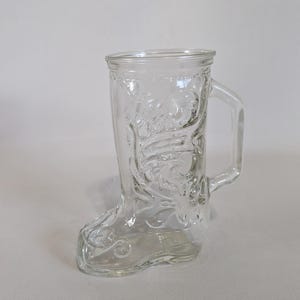 May include: A clear glass boot-shaped mug with a handle. The mug features a detailed design of a cowboy boot.