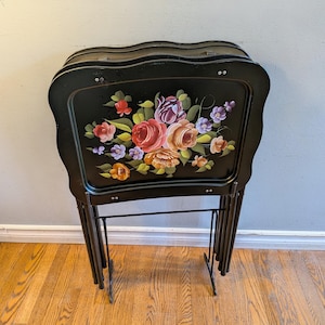 May include: A set of black folding snack trays with a scalloped edge design. The top tray features a hand-painted floral arrangement with red, orange, purple, and pink roses and green leaves against a glossy black background.