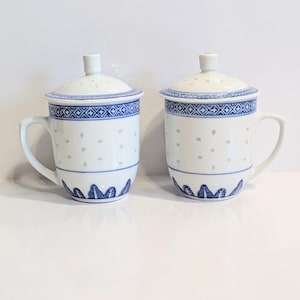 Op de afbeelding: Twee witte keramische theemokken met deksels. Elke mok heeft een blauw geometrisch patroon rond de rand en de basis, en een stippenpatroon op de body. De deksels hebben een kleine knop en een geperforeerd ontwerp.