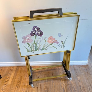 May include: A set of four rectangular TV tray tables with a gold-tone metal frame and dark brown wooden accents. Each tray features a light-colored surface decorated with a floral illustration of purple and pink irises and a small blue butterfly.