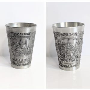 May include: Two pewter-coloured drinking glasses. Each glass has a detailed embossed design of a townscape with buildings and text. The glasses are cylindrical with a slightly flared rim and a detailed base.