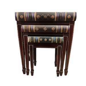 May include: Set of three nesting tables with faux book tops. The tops are brown with gold accents and the spines are decorated with gold lettering. The tables are made of dark wood with gold accents.