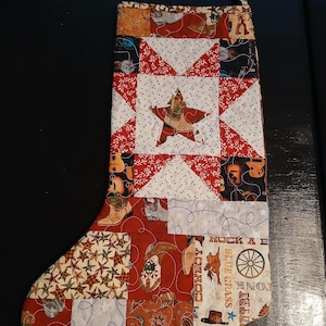 May include: A quilted Christmas stocking featuring a patchwork design with various western-themed fabrics. Patterns include cowboy boots, guitars, stars, paisley, and text reading "COWBOY" and "BLUE GRASS". The center features a large star motif.