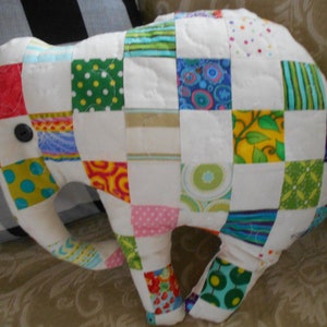 May include: A patchwork elephant toy made of colourful fabric squares. The elephant is white with a black button eye and has a variety of patterns including polka dots, stripes, and floral designs.