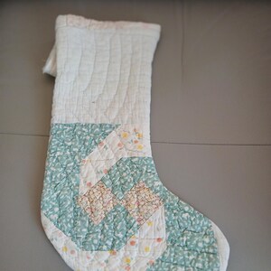 May include: A quilted Christmas stocking featuring a patchwork design with white, sage green, and floral patterned fabric squares. The stocking has a vertical quilted texture throughout and a folded cuff at the top, showcasing a soft, stitched construction.