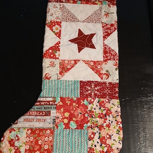 May include: A quilted Christmas stocking featuring a patchwork design with red, white, and teal fabrics. The upper section displays a central red star on a white background, while the lower portion includes floral patterns, zigzags, and text.