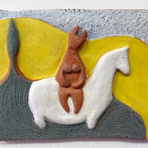 Op de afbeelding: Een keramisch reliëfsculptuur met een bruine figuur die op een wit paard rijdt tegen een gele en grijze achtergrond. De achtergrond toont gestileerde heuvels en een getextureerde lucht. Het kunstwerk is een reliëf.