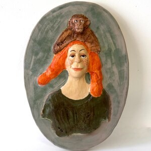 May include: A ceramic wall plaque depicting a woman with vibrant red hair and a monkey resting on her head. The woman has freckles and wears a dark green top. The background is an oval in shades of green.