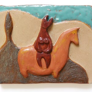 Op de afbeelding: Een keramische tegel met een bruin figuur dat een lichtoranje paard berijdt. De achtergrond is lichtbruin met een blauwe lucht.