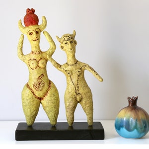 May include: Two yellow ceramic figurines of a woman and a man with horns, standing on a black base. The woman is wearing a red crown and the man is holding his hand up. A blue and brown ceramic pomegranate sits on a white surface.