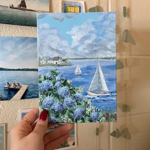 Op de afbeelding: Een kleine schildering toont een kustlandschap met een zeilboot op blauw water, witte wolken en blauwe bloemen. Witte huizen zijn zichtbaar aan de kust. De schildering wordt door een persoon vastgehouden.
