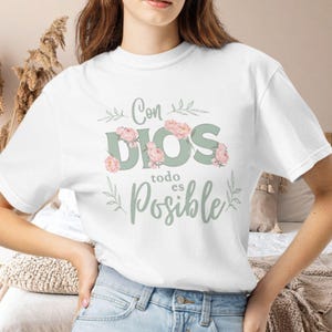 May include: A white short-sleeved t-shirt featuring the text Con DIOS todo es Posible in sage green lettering. The design is decorated with soft pink floral accents and delicate sage green leaf illustrations surrounding the words.