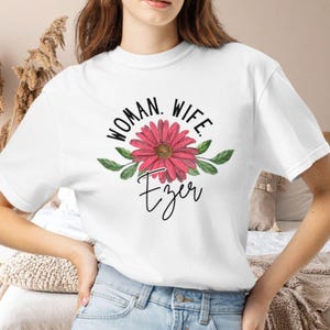 May include: A white short-sleeved t-shirt featuring a graphic print of a pink daisy flower with green leaves. Above the flower, the text reads WOMAN. WIFE. in black capital letters, and below the flower, the word Ezer is written in black script.