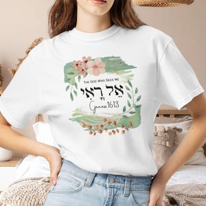 May include: A white short-sleeved t-shirt featuring a graphic print with green watercolor brushstrokes, pink flowers, and brown foliage. The text reads The God Who Sees Me, the Hebrew characters El Roi, and the scripture reference Genesis 16:13.
