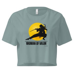 May include: A light blue crop top with a graphic of a woman in a fighting stance holding a sword in front of a yellow circle. The text "WOMAN OF VALOR" is printed below the graphic.
