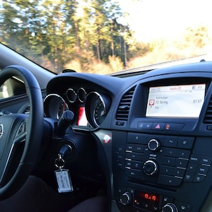 Può includere: Interno auto con cruscotto e volante. Il volante presenta il logo Opel. Il cruscotto è grigio scuro con un display centrale che mostra un'interfaccia radio. L'auto è in movimento, con uno sfondo sfocato di alberi visibile attraverso il parabrezza.