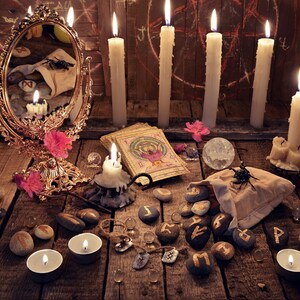May include: A mystical scene featuring a gold-framed mirror, lit candles, tarot cards, and rune stones on a wooden table. Pink flowers and a crystal ball add to the esoteric ambiance. A small bag of rune stones is open, with some stones scattered around.