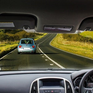 Può includere: Vista dall'interno di un'auto, che mostra una strada tortuosa con un'auto azzurra in lontananza. Cruscotto, volante e parasole sono visibili. Paesaggio verde.