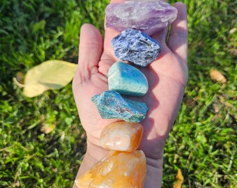 Può includere: Una mano che tiene una collezione di pietre preziose colorate e lucide. Le pietre includono quarzo trasparente, ametista, sodalite, amazzonite, calcite arancione e corniola. Le pietre sono disposte verticalmente su uno sfondo di erba verde.