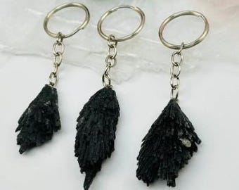 Può includere: Tre portachiavi in kianite nera con anelli e catene in metallo color argento. I cristalli di kianite hanno un aspetto ruvido e strutturato. I portachiavi sono esposti su una superficie bianca con uno sfondo sfocato.