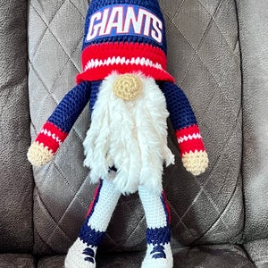 May include: A crocheted gnome wearing a blue and red hat with the word "GIANTS" on it. The gnome is sitting on a gray couch.