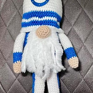 May include: A handmade crochet gnome in the colors of the Indianapolis Colts football team. The gnome has a white hat with a blue horseshoe logo, a white beard, and blue and white striped legs and arms. The gnome is wearing white shoes with blue laces.