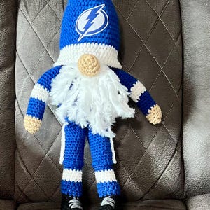 May include: A crocheted gnome wearing a blue and white hat with the Tampa Bay Lightning logo. The gnome is wearing a blue and white crocheted outfit with white trim and black shoes.