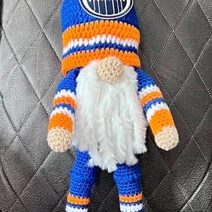 May include: A handmade gnome doll wearing a blue and orange knit hat with the Oilers logo. The gnome has a white beard, tan nose, and is dressed in blue and orange striped clothing and black shoes.