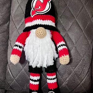 May include: A handmade crochet gnome doll in the colors of the New Jersey Devils hockey team. The gnome has a black hat with the team logo, a white beard, and red and black striped arms and legs. The gnome is wearing black skates.