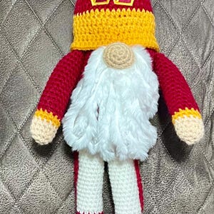 May include: A handcrafted crochet gnome doll featuring a tall red pointed hat with a yellow band and a burgundy and gold W logo. The gnome has a white faux fur beard, a beige nose, red arms with yellow cuffs, and white legs with red stripes.