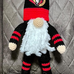 May include: A crocheted gnome doll featuring a black and red pointed hat with an Ottawa Senators logo patch. The doll has a long white faux fur beard, a beige nose, and black and red striped arms and legs. It wears black skates with white laces.