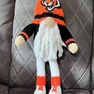 May include: A crocheted gnome wearing a hat with a Cincinnati Bengals logo. The gnome is wearing black and orange striped clothing and white shoes with orange accents.