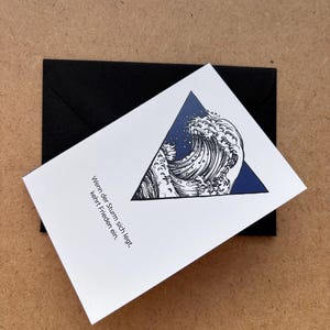 May include: A white greeting card with a blue triangle design featuring a wave illustration, resting on a black envelope. The card has German text. The background is a light brown surface.