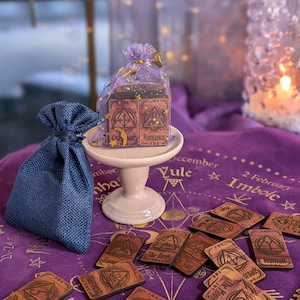 May include: A set of wooden oracle cards with a purple background and gold lettering. The cards are arranged in a circle around a small, pink pedestal. The pedestal holds a small, purple bag with a gold drawstring. The bag contains a set of wooden oracle cards. The cards are labeled with words like "The Scythe", "Romance", "Harvest", "The Birds", "The Moon", "Woman", and "Woods Will Guide The Ride".