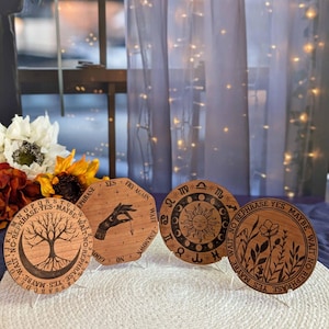 May include: Three wooden circular divination boards with different designs. The first board features a tree with roots and branches, a crescent moon, and the words "YES TRY AGAIN WAIT NO REPHRASE YES MAYBE WAIT NO REPHRASE". The second board features a hand holding a pendulum and the words "YES TRY AGAIN WAIT UNKNOWN ON". The third board features a sun with a face, a crescent moon, and the words "NO REPHRASE YES MAYBE WAIT NO REPHRASE YES MAYBE WAIT NO REPHRASE".