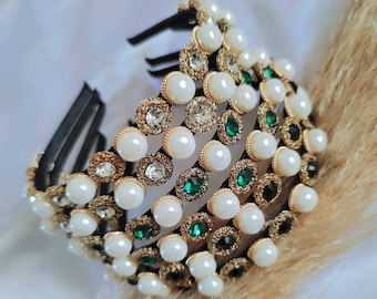 Puede incluir: Una diadema dorada con varias filas de perlas falsas y piedras verdes. La diadema está hecha de un material elástico negro.
