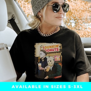 May include: Black t-shirt with a graphic of Michael Myers and Jason Voorhees holding coffee cups in front of a Dunkin' Donuts store. The text "Dunkin' Donuts" is on the sign above the store.