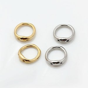 May include: Two gold-toned and two silver-toned metal rings with an irregular, organic shape. Each ring features a slightly flattened section on one side with two small, circular indentations or holes, creating a distinct, textured appearance.
